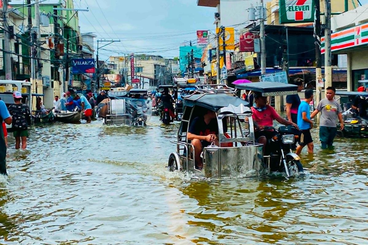 A tricycle pushes through deep floodwater along a commercial street in Apalit while residents wade past the shopfronts.