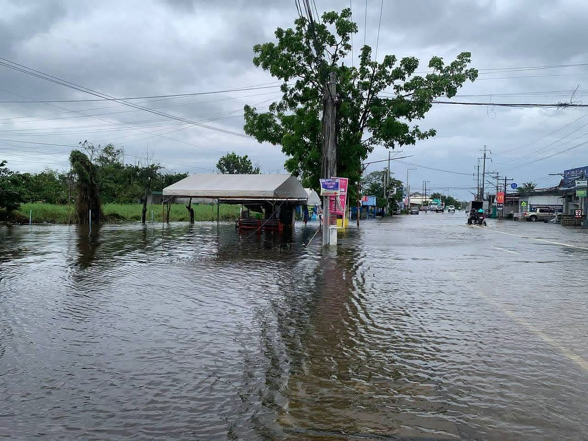 Floodwater covers a main roadside and reaches businesses and a waiting shed in San Simon under an overcast sky.